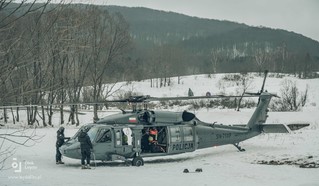 Polowe lądowisko śmigłowców przed dziobem stojącej na pokrytej śniegiem polanie dwóch crew chiefów asekurujących moment uruchomienia silników maszyny. W tle bujny zimowy las na pobliskich wzniesieniach.