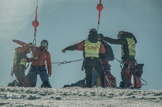 Stojąc na pokrytym śniegiem wzniesieniu trzej uczestnicy ćwiczeń pod okiem dwóch instruktorów GOPR-u przypinają się karabińczykami uprzęży asekuracyjnych do zwisających z pokładu Black Hawka lin wyciągarki pokładowej.