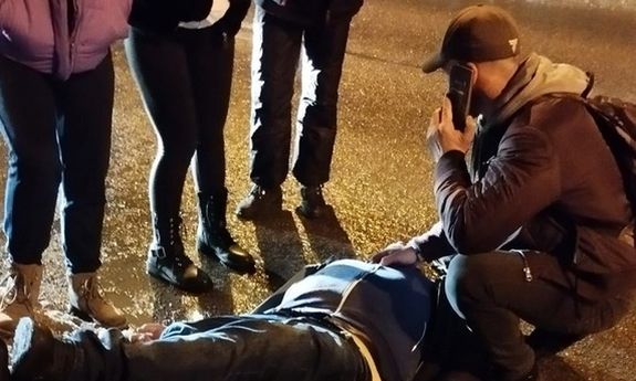mężczyzna klęczy przy leżącej osobie i rozmawia przez telefon, obok widać nogi trzech stojących osób
