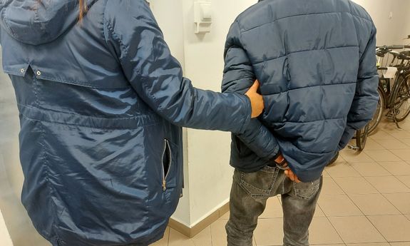 Policjantka trzyma za ramię zatrzymanego mężczyznę, który ręce ma skute kajdankami - widok z tyłu