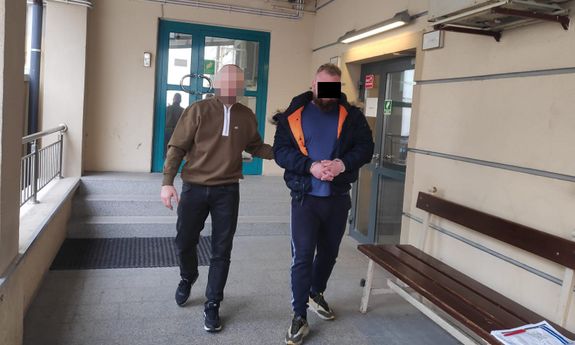 policjant prowadzi zatrzymanego, który ma ręce skute kajdankami