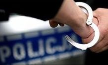 ręka osoby w trakcie zakuwania w kajdanki, z tyłu napis policja na niebieskim tle