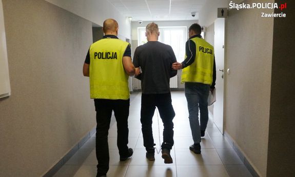Na zdjęciu widoczni dwaj policjanci ubrani w kamizelki odblaskowe z napisem POLICJA, którzy prowadzą korytarzem sprawcę rozboju. Mężczyzna ma kajdanki zespolone założone na ręce i na nogi