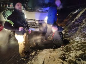 zatrzymany mężczyzna w kajdankach siedzi na ziemi, obok niego stoją funkcjonariusze, w tle skradziony pojazd