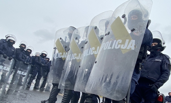na zdjęciu policjanci z uniesionymi tarczami z napisem policja. stoją w szeregu czteroosobowym. w tle inni policjanci