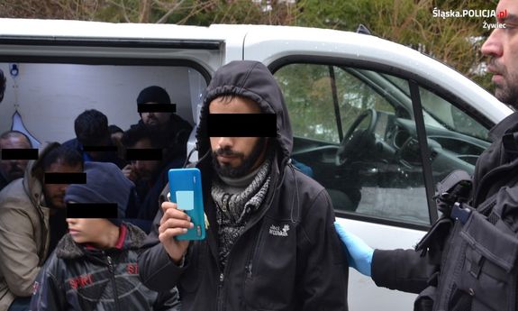 zdjęcie kolorowe widać otwarte drzwi samochodu w środku siedzą osoby obok fragment policjanta