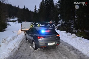 zdjęcie kolorowe zrobione na drodze w lesie na pierwszym planie zatrzymany samochód w tle widać radiowóz z włączonymi sygnałami świetlnymi obok samochodu stoją policjanci