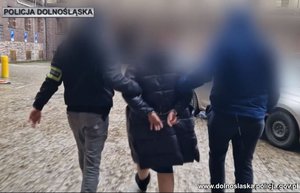 policjanci z opaskami na rękach prowadzą zatrzymaną osobę do samochodu