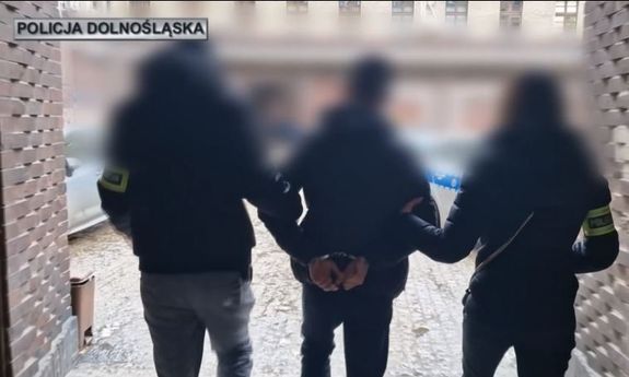 policjanci z opaskami na rękach prowadzą zatrzymaną osobę do samochodu