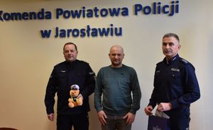 Pomiędzy policjantami stoi mężczyzna. Policjant znajdujący się po lewej stronie trzyma w ręce misia policyjnego