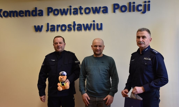 Pomiędzy policjantami stoi mężczyzna. Policjant znajdujący się po lewej stronie trzyma w ręce misia policyjnego