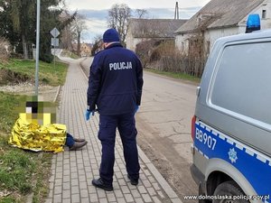 Na zdjęciu policjant stojący przy radiowozie pochylający się nad mężczyzną w kocu termicznym siedzącym na chodniku