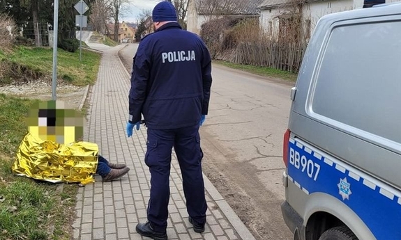 Na zdjęciu policjant stojący przy radiowozie pochylający się nad mężczyzną w kocu termicznym siedzącym na chodniku
