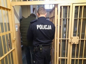 policjant prowadzi zatrzymanego - z boku widoczne są kraty