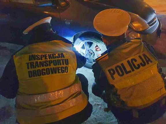 pora nocna, policjant i inspektor transportu drogowego kucają i sprawdzają ogumienie samochodu - widok z tyłu