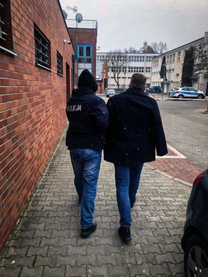 Zdjęcie przedstawia umundurowanego policjanta, który prowadzi po swojej prawej stronie zatrzymanego mężczyznę.