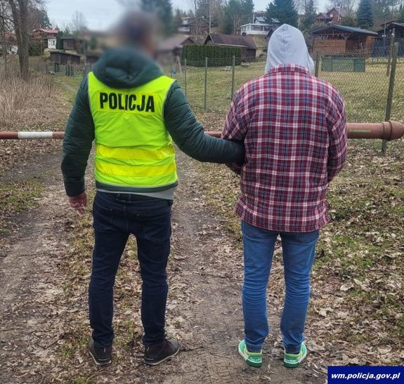 Zdjęcie przedstawia policjanta ubranego w odblaskową kamizelkę z czarnym napisem POLICJA na plecach, który przytrzymuje za lewe przedramię zatrzymanego mężczyznę. Mężczyzna ten ma założony ma głowę szary kaptur, ubrany jest w kurtkę bordową w jasną kratę, niebieskie spodnie jeasowe i jasne buty sportowe. Obaj stoją tyłem do obiektywu aparatu.