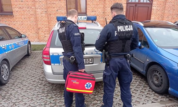 dwaj policjanci z torbą medyczną stoją przy bagażniku radiowozu - widok z tyłu