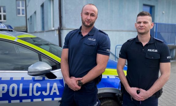 Dzielnicowi mł. asp. Bartosz Gorczyca i mł. asp. Krystian Biela na tle oznakowanego radiowozu i budynku komendy