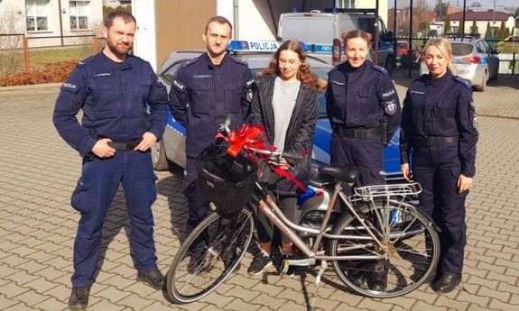 Dwóch policjantów w mundurach i dwie policjantki stoją obok młodej dziewczyny, która stoi w środku, przed nimi ustawiony rower z czerwoną kokardą, z tyłu stoją zaparkowane radiowozy