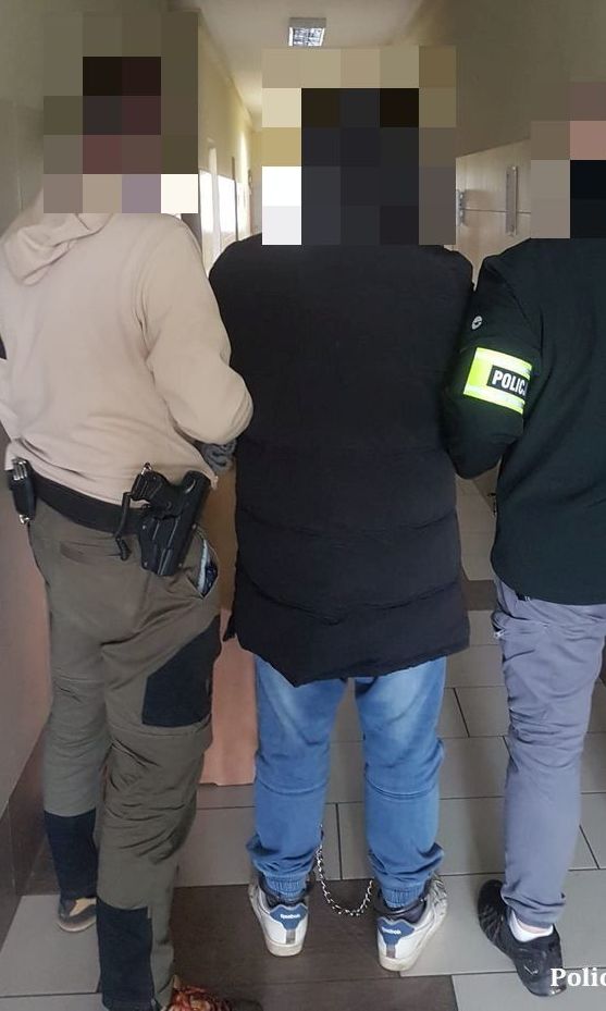 Zdjęcie przedstawia dwóch nieumundurowanych policjantów, którzy prowadzą pomiędzy nimi zatrzymanego, który ubrany jest w ciemną długą kurtkę i niebieskie spodnie jeansowe. Wszyscy stoją tyłem do zdjęcia.