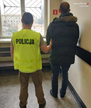 Policjant z zatrzymanym stoi przed zakratowanym oknem