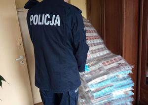 Zdjęcie przedstawia sylwetkę umundurowanego policjanta, który stoi przed stosem ułożonych opakowań z podrobionymi papierosami.