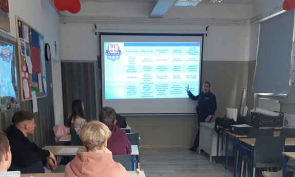 policjantka w klasie lekcyjnej, stoi przed ekranem, na którym wyświetlana jest prezentacja, w ławkach siedzą dzieci