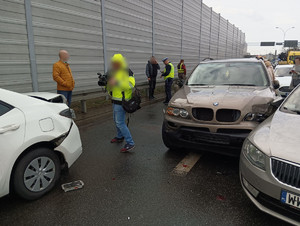 zdjęcie z sytuacji na drodze, uszkodzone samochody, wokół stojące osoby oraz osoba z kamerą