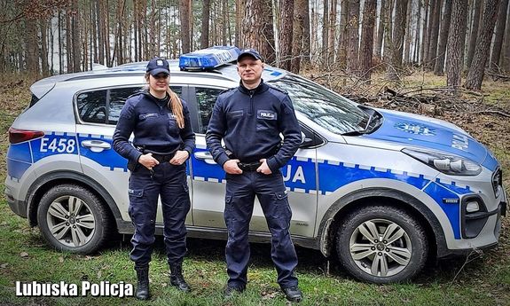 Zdjęcie przedstawia umundurowaną policjantkę i umundurowanego policjanta, którzy stoją na tle oznakowanego radiowozu.