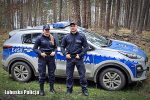 Zdjęcie przedstawia umundurowaną policjantkę i umundurowanego policjanta, którzy stoją na tle oznakowanego radiowozu.