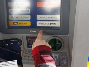 Zdjęcie przedstawia dłoń sięgającą do klawiatury bankomatu na ekranie którego wyświetla się kilka komunikatów.