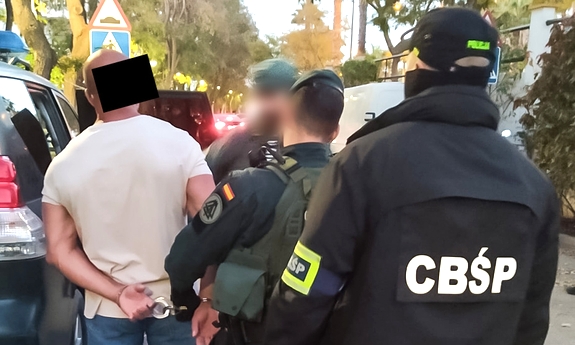 funkcjonariusz CBŚP i funkcjonariusze CBŚP, Guardia Civil z zatrzymanym mężczyzną zakutym w kajdanki przy radiowozie