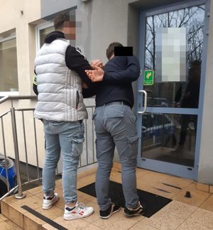 Zdjęcie przedstawia nieumundurowanego policjanta, który wprowadza do budynku zatrzymanego mężczyznę. Mężczyzna ten ma kajdanki otwarte na rękach trzymanych z tyłu.