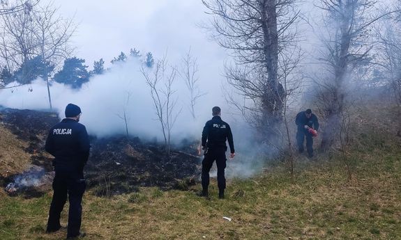 trzej policjanci na skraju palącej się łąki, jeden trzyma w ręku gaśnicę