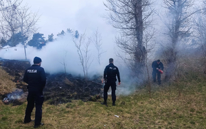 trzej policjanci na skraju palącej się łąki, jeden trzyma w ręku gaśnicę