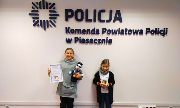 dwie dziewczynki stojące na tle napisu Komenda Powiatowa Policji w Piasecznie, w rękach trzymają nagrody