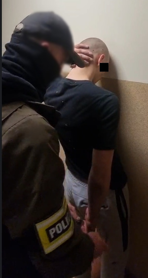 Zdjęcie przedstawia policjanta ubranego po cywilnemu, który ma na prawym przedramieniu odblaskową opaskę i zakłada kajdanki na ręce trzymane z tyłu opartemu o ścianę mężczyźnie. Część twarzy tego mężczyzny zasłania czarny prostokąt.