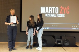 Zdjęcie przedstawia umundurowaną policjantkę, która trzyma w rękach kartki. Obok niej, po prawej stronie zdjęcia stoją dwie młode dziewczyny.