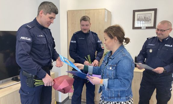 Zdjęcie przedstawia umundurowanych policjantów, którzy stoją w jednym pomieszczeniu. Przed jednym z nich stoi kobieta i trzyma w dłoniach niebieską kartkę.