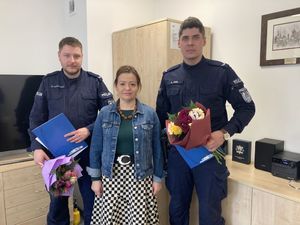 Zdjęcie przedstawia umundurowanych policjantów, którzy stoją w pomieszczeniu. Trzymają w dłoniach kolorowy bukiet kwiatów i niebieską kartkę. Przed nimi stoi kobieta.