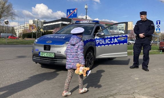 Zdjęcie przedstawia małą dziewczynkę w policyjnej czapce, która podchodzi do oznakowanego radiowozu.