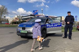 Zdjęcie przedstawia małą dziewczynkę w policyjnej czapce, która podchodzi do umundurowanych policjantów, którzy stoją przy oznakowanym radiowozie.