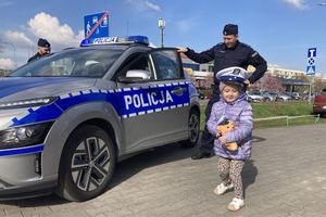 Zdjęcie przedstawia dziewczynkę stojącą przy radiowozie z umundurowanym policjantem.