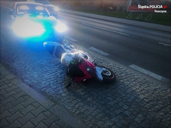 motocykl, który leży na ziemi, za nim zaparkowany samochód osobowy z włączonymi sygnałami świetlnymi