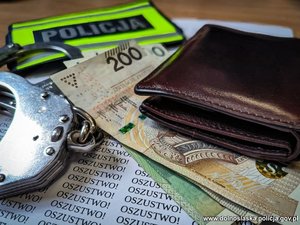 Na powierzchni leżące pieniądze, portfel, kajdanki oraz opaska z napisem Policja, na spodzie kartka z napisami Oszustwo!
