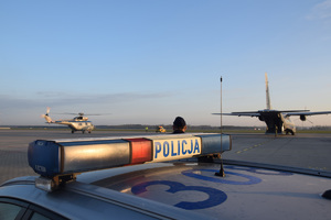 Sygnały świetlne na dachu policyjnego radiowozu, w tle samolot Casa i śmigłowiec Sokół na lotnisku.