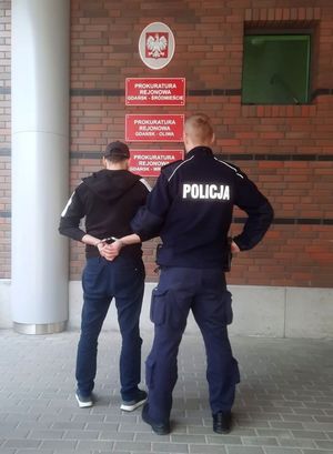 Policjant z zatrzymanym stoi przed budynkiem prokuratury. Zatrzymany  na rękach trzymanych z tyłu ma załozone kajdanki. - widok z tyłu