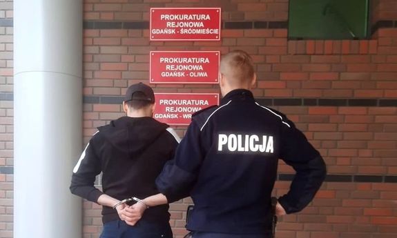 Policjant z zatrzymanym stoi przed budynkiem prokuratury. Zatrzymany  na rękach trzymanych z tyłu ma załozone kajdanki. - widok z tyłu