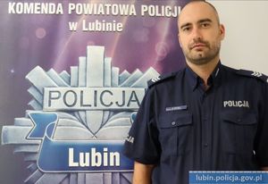 umundurowany policjant stoi na tle ścianki z napisem Komenda Powiatowa Policji w Lubinie